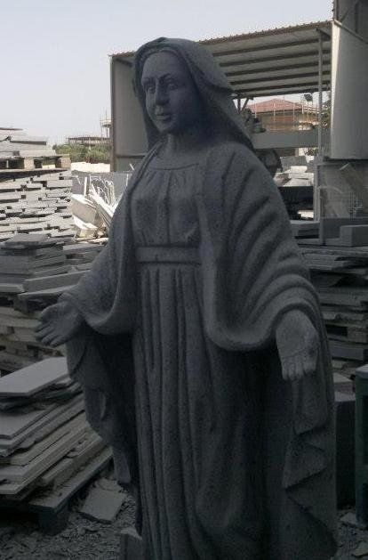 Statua in pietra raffigurante una donna con le braccia tese e abiti drappeggiati, probabilmente di carattere religioso. In piedi vicino a pile di lastre di pietra.