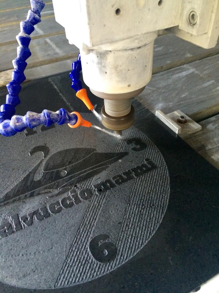 Logo e numeri incisi con macchina CNC su pietra scura. Sono visibili i tubi blu del refrigerante.