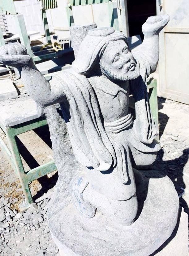 Scultura in pietra raffigurante un uomo inginocchiato con le braccia alzate, che indossa un turbante e una tunica.