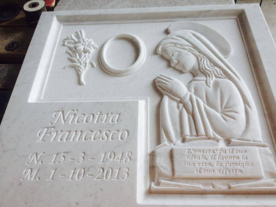 Monumento commemorativo in marmo con motivo floreale scolpito, cerchio vuoto, donna in preghiera e nomi e date incisi.
