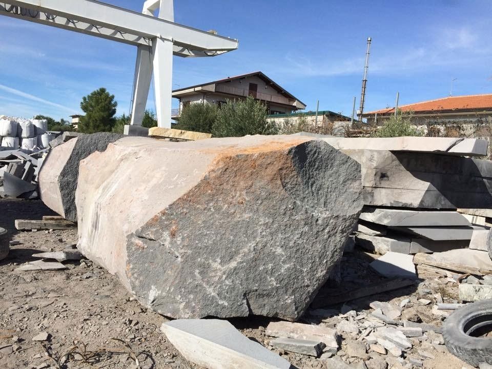 Grande blocco di granito tagliato all'esterno, tonalità di grigio e arancione, con una gru a portale ed edifici sullo sfondo.