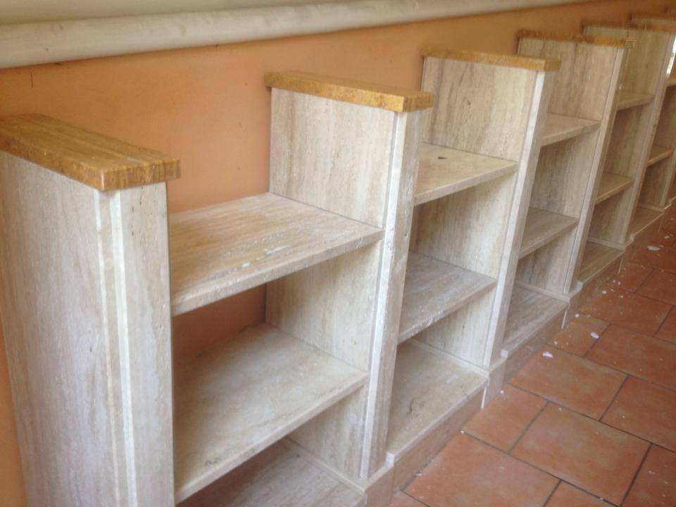 Fila di librerie in legno chiaro contro una parete arancione, su un pavimento in piastrelle di terracotta.