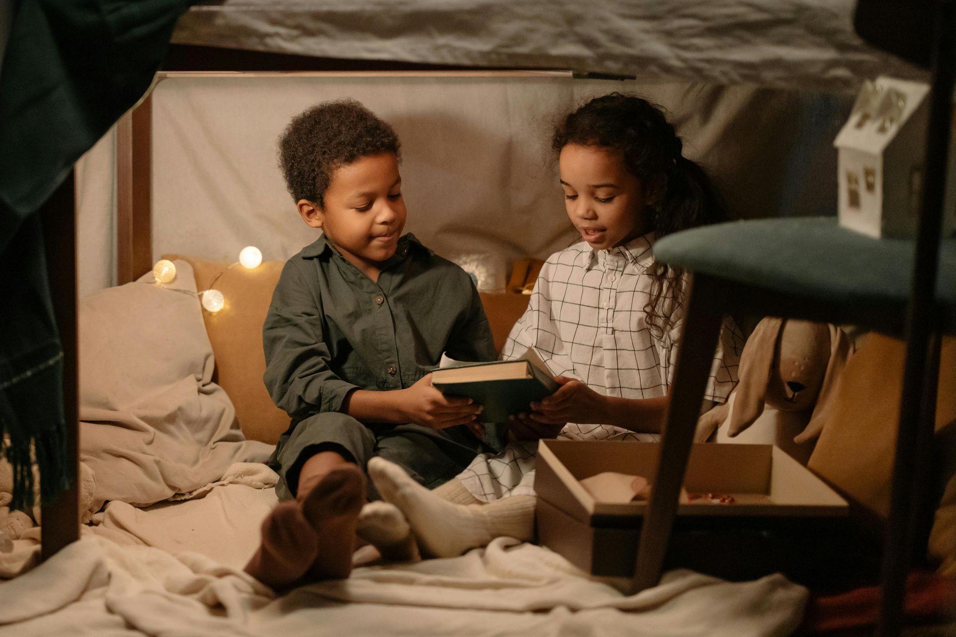 Two children looking at a book inside a blanket fort, lit with fairy lights.