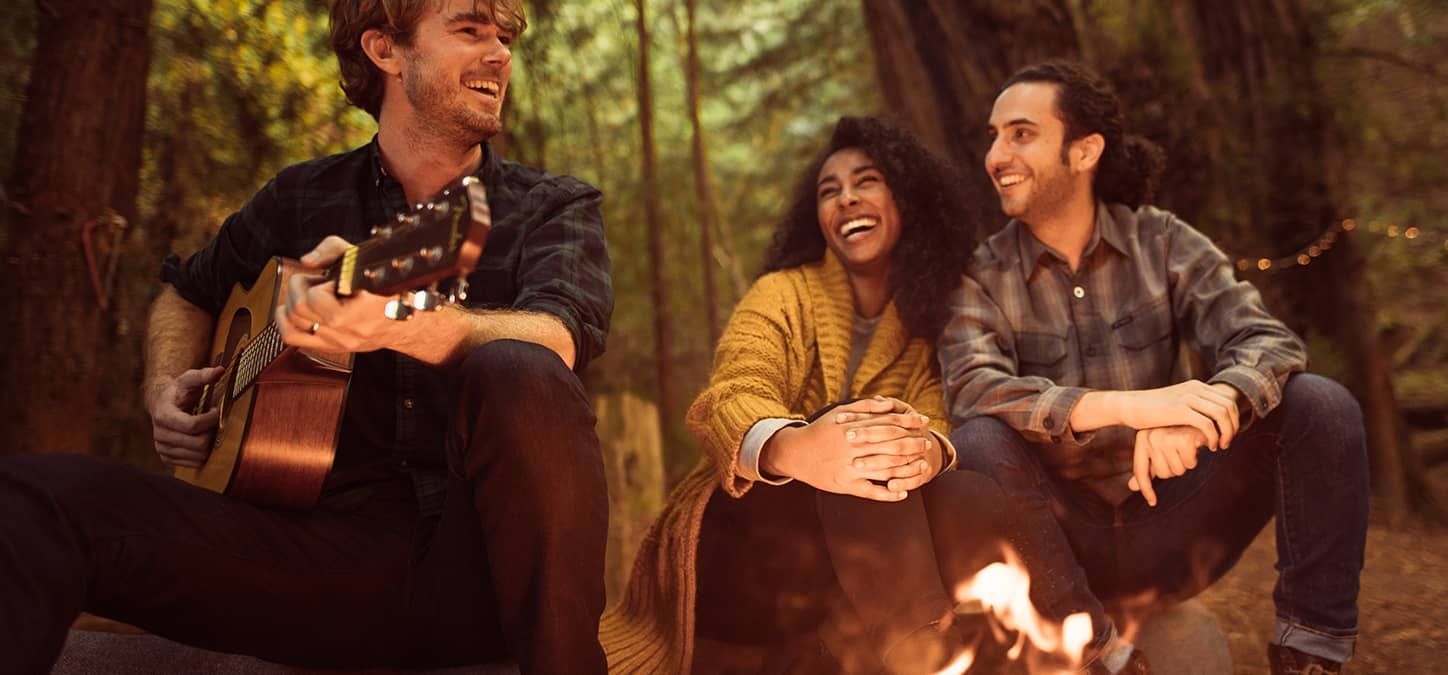 A group of people are sitting playing guitar around a campfire in the woods.