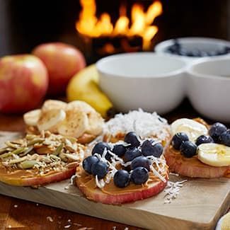 Apple slices topped with peanut butter and various fruits and seeds on a wooden board.