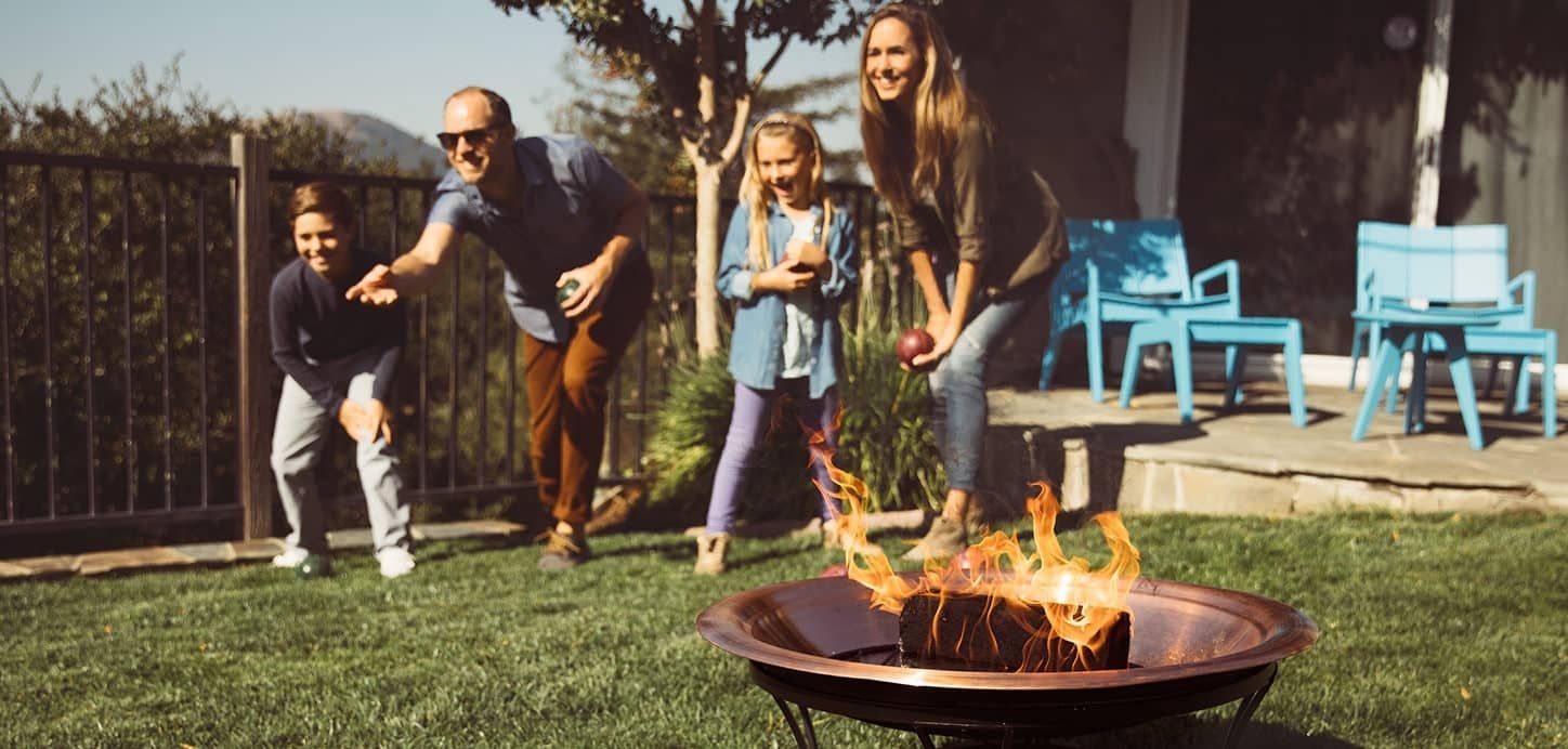 A family is playing bocce on backyard lawn with a duraflame indoor/outdoor firelog burning in fire pit in foreground