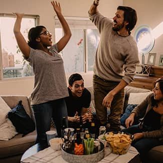Four people celebrate in a living room, cheering and gesturing with excitement around a table with snacks and drinks.