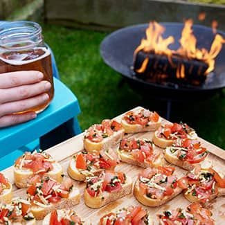 Bruschetta appetizers on wooden board, beer in hand, fire pit with flames in background.