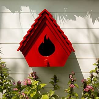 Red, DURAFLAME flame-shaped birdhouse with a large black opening, mounted on a white wall with foliage below.