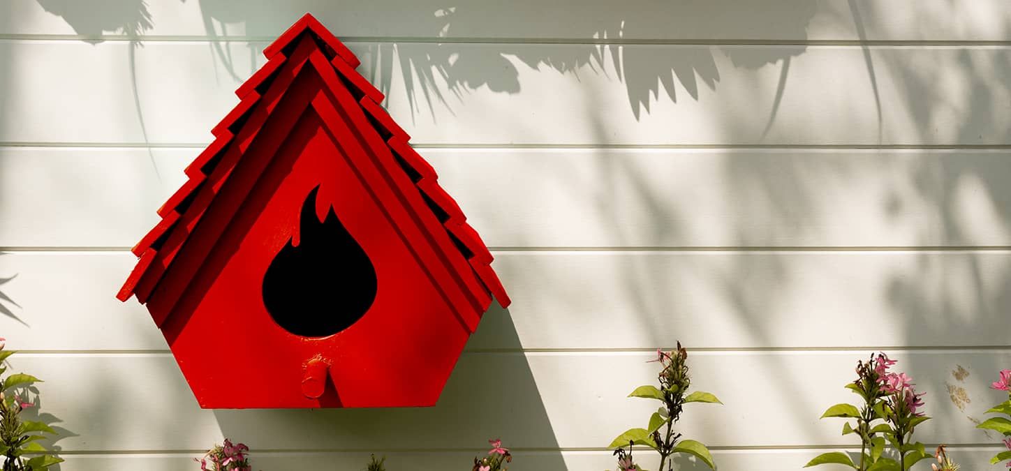 A red birdhouse whose opening is in the shape of a duraflame flame logo is hanging on a white wall.