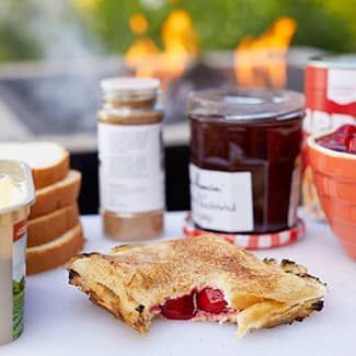 Cherry campfire pie with jam, spice, bread, and butter on a table near a grill and fire.