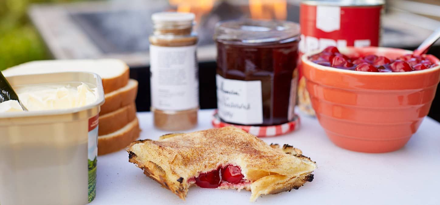 A campfire pie sandwich is sitting on a table next to a bowl of berries and a jar of jam in front of a fire pit