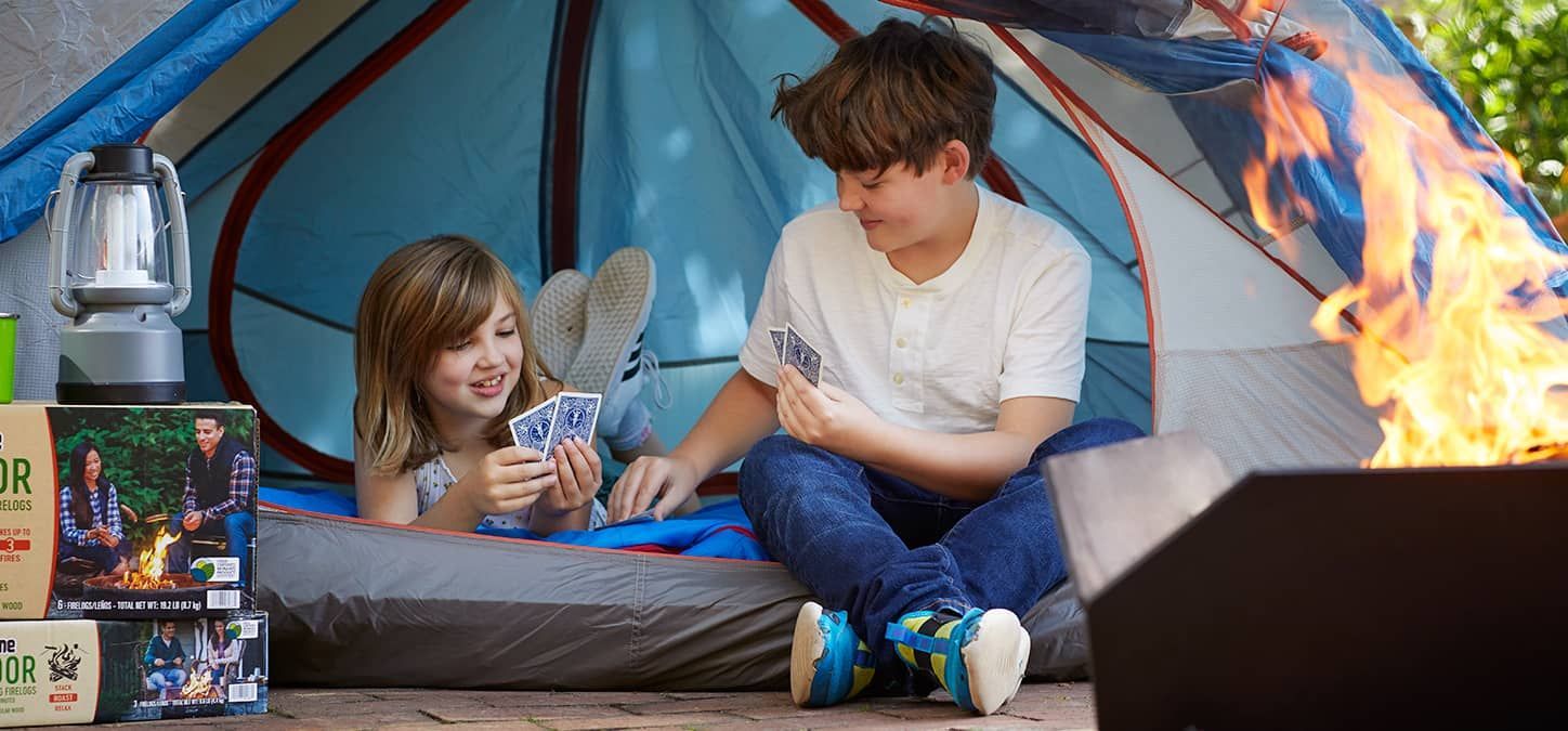 A boy and a girl are playing cards in a tent next to a fire.
