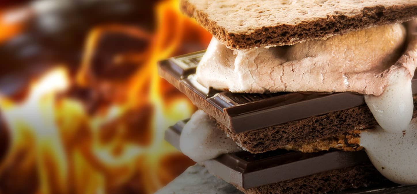 A chocolate bomb s'mores  is stacked with marshmallows, chocolate and chocolate graham crackers in front of a fire.