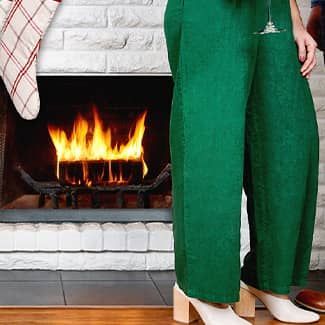 A woman in green pants is standing in front of a fireplace holding a glass of wine.