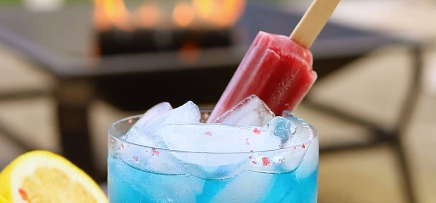 A blue crackletini drink with ice and a red popsicle sticking out of it, with a fire pit in the background