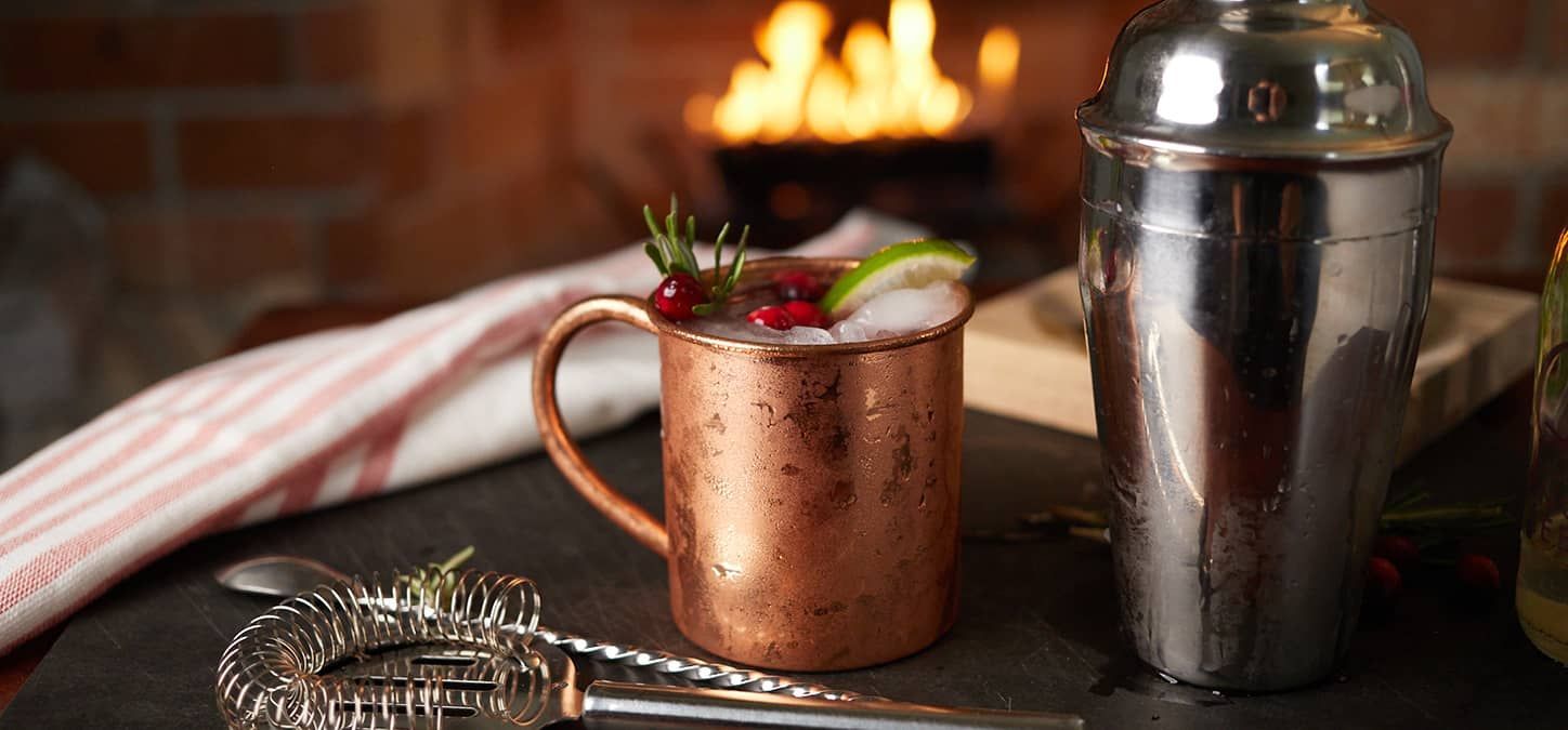 A copper mug filled with a cranberry moscow mule next to a shaker on a table in front of a fireplace