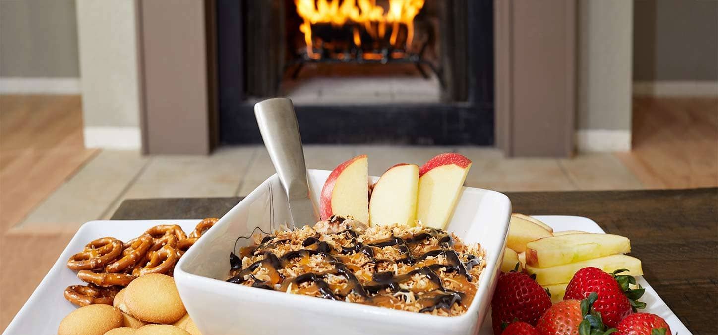 A bowl of cookie dip with fruit and pretzels on a plate in front of a fireplace.