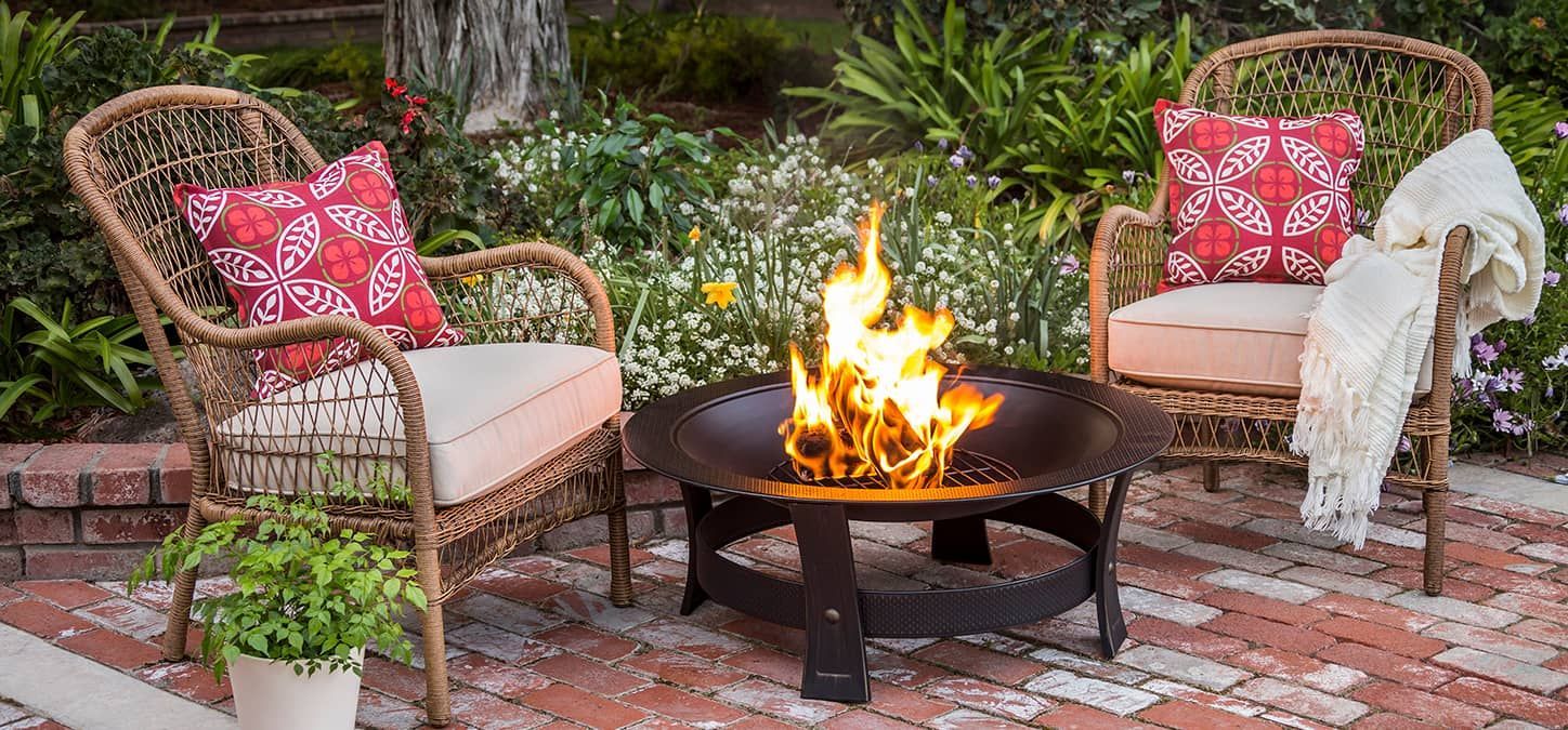 A fire pit is sitting on a brick patio next to two chairs with pillows