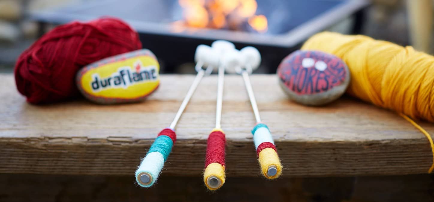 Three marshmallow rosting sticks decorated with yarn are sitting on a wooden table next to a fire pit