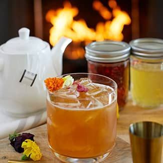 Earl grey tea cocktail with ice, flower garnish, teapot, jars of ingredients, and fire in background.