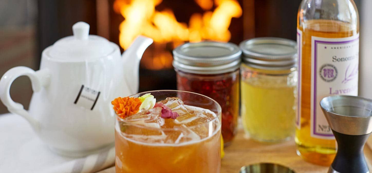 An earl grey cocktail with garnishes, tea pot, jars of ingredients, and lavender syrup. Fireplace in background.