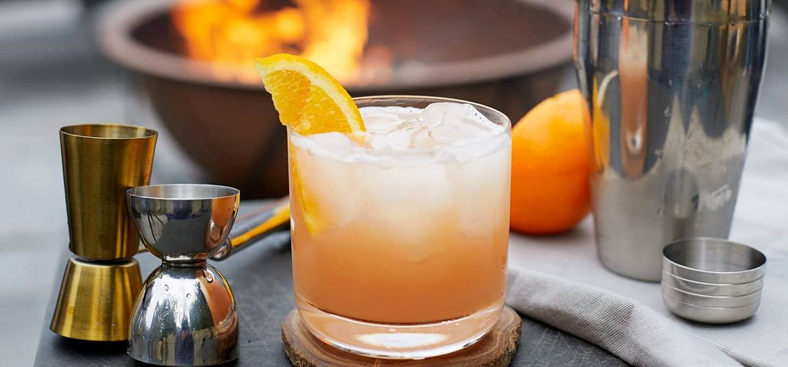 Elderflower greyhound Cocktail with orange garnish on a table, beside a jigger, shaker, and napkin near a firelit background