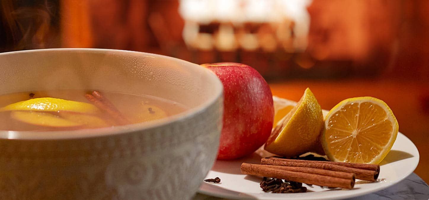 A bowl of mulled wine next to a plate of fruit and cinnamon sticks with fire burning in the background