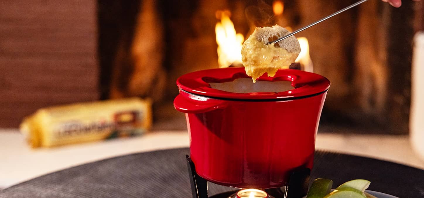 A person is dipping a piece of bread into cheese in a red fondue pot in front of the fire with a Crackleflame firelog