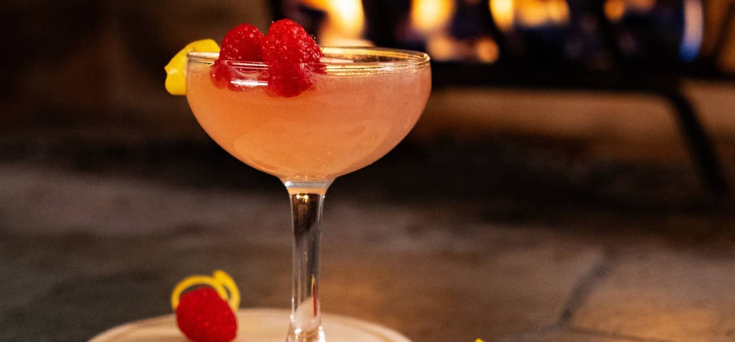 A French 75 cocktail in a martini glass with raspberries on top in front of a fire