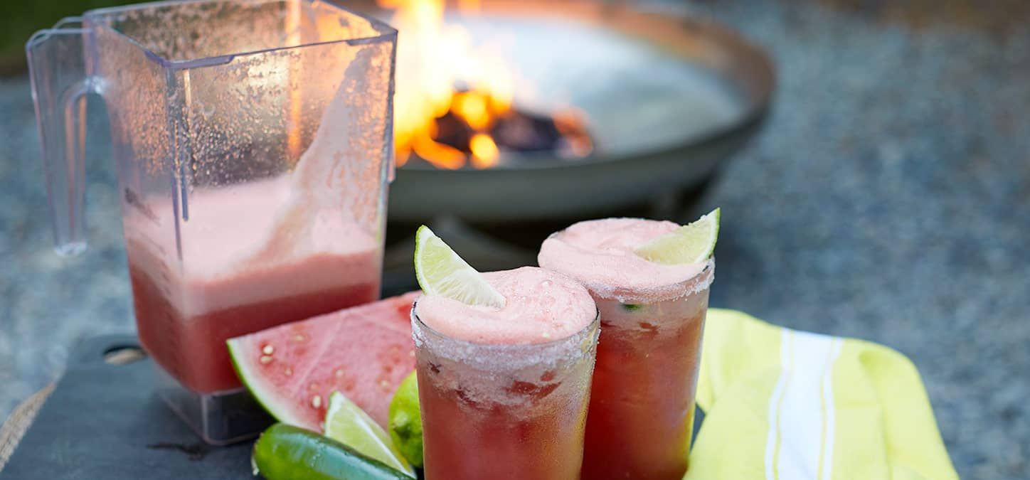 Two glasses of watermelon margarita next to a pitcher of watermelon juice with a fire pit in the background