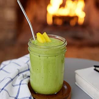 A green smoothie in a mason jar with a straw and pineapple slices on a table in front of a fire