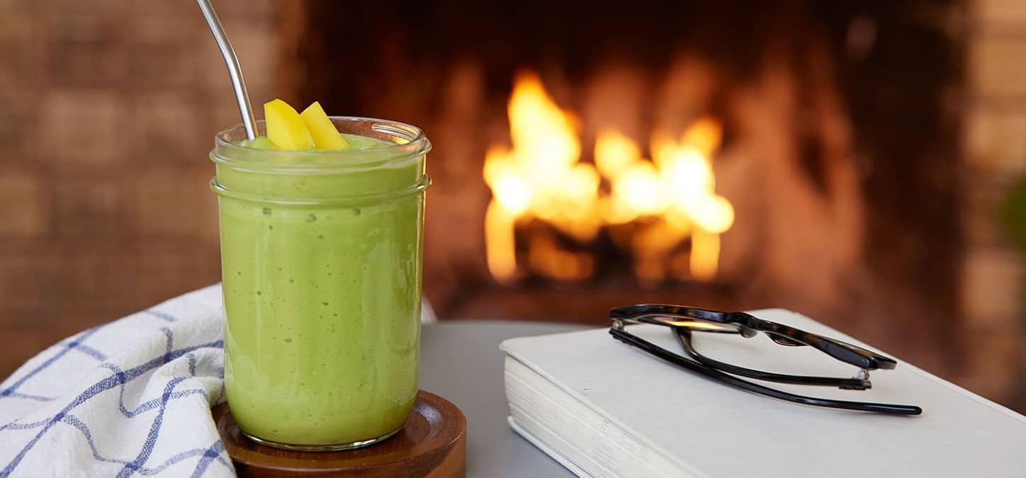 A green smoothie is sitting on a table next to a book and glasses in front of a fireplace