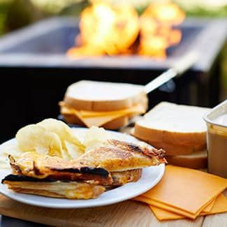 Grilled cheese sandwich on a plate with chips, cheese slices, and a fire pit in the background.