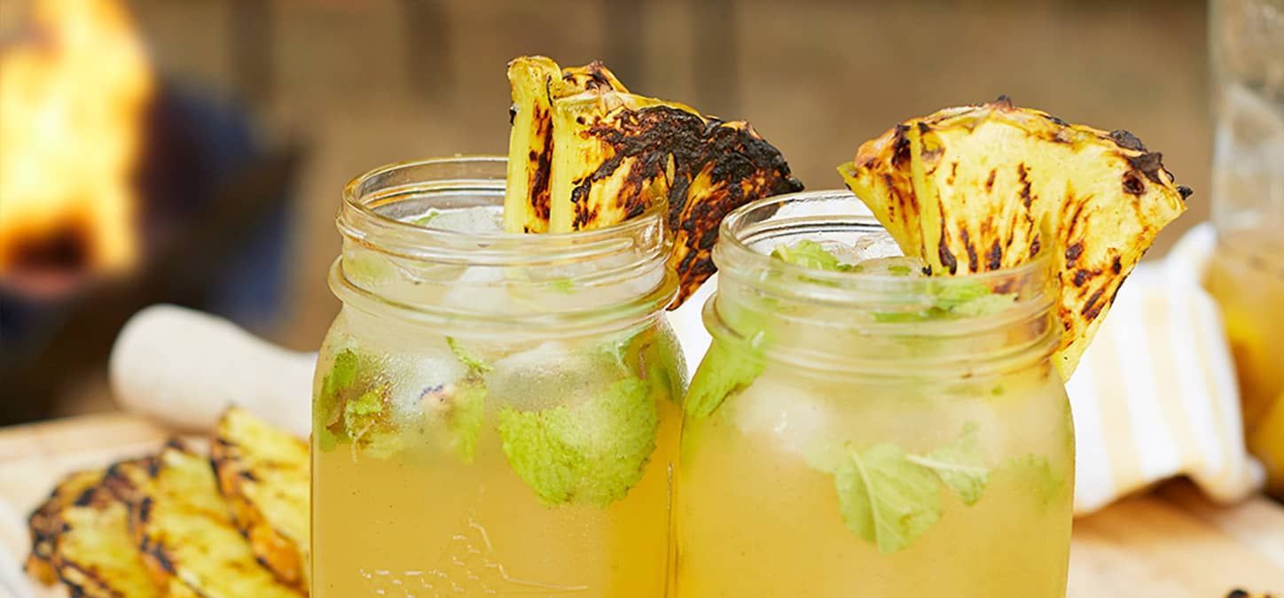 Two mason jars filled with pinapple mojito garnished with grilled pineapple slices on a table.
