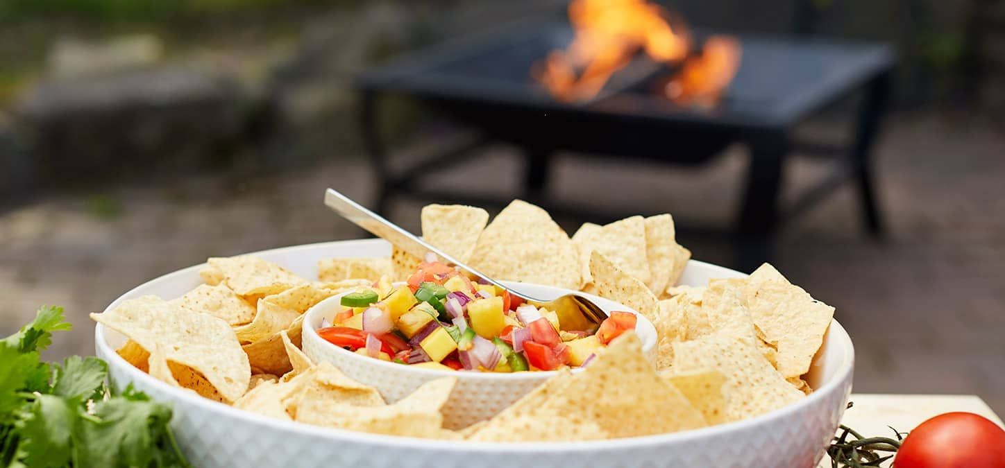 A bowl of chips and salsa with a spoon in it is on a table in front of a fire pit.