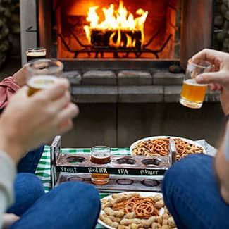 People toasting with beer flights near a fireplace, accompanied by snacks.