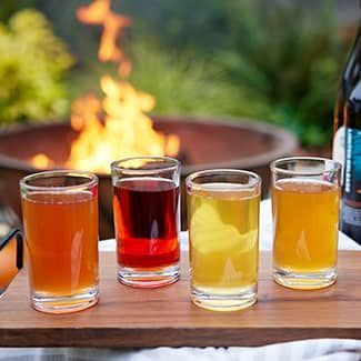 Four shot glasses with different colored drinks on a wooden board; fire pit in background.