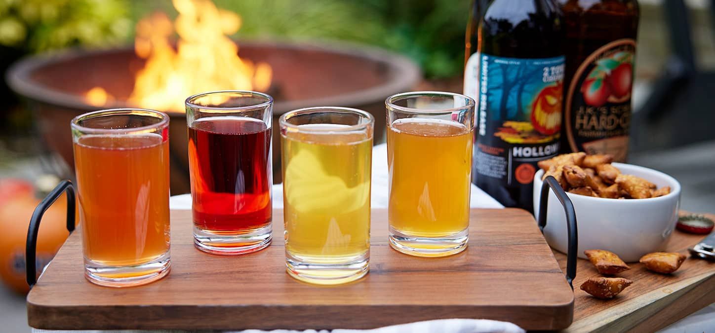A wooden tray with four glasses of different types of cider on it.