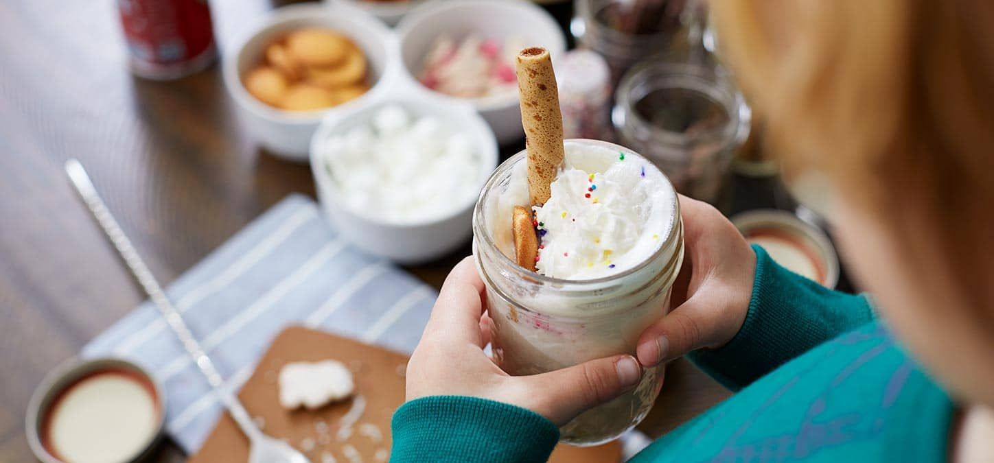 A person is holding a cocoa in a jar with whipped cream and cookies.