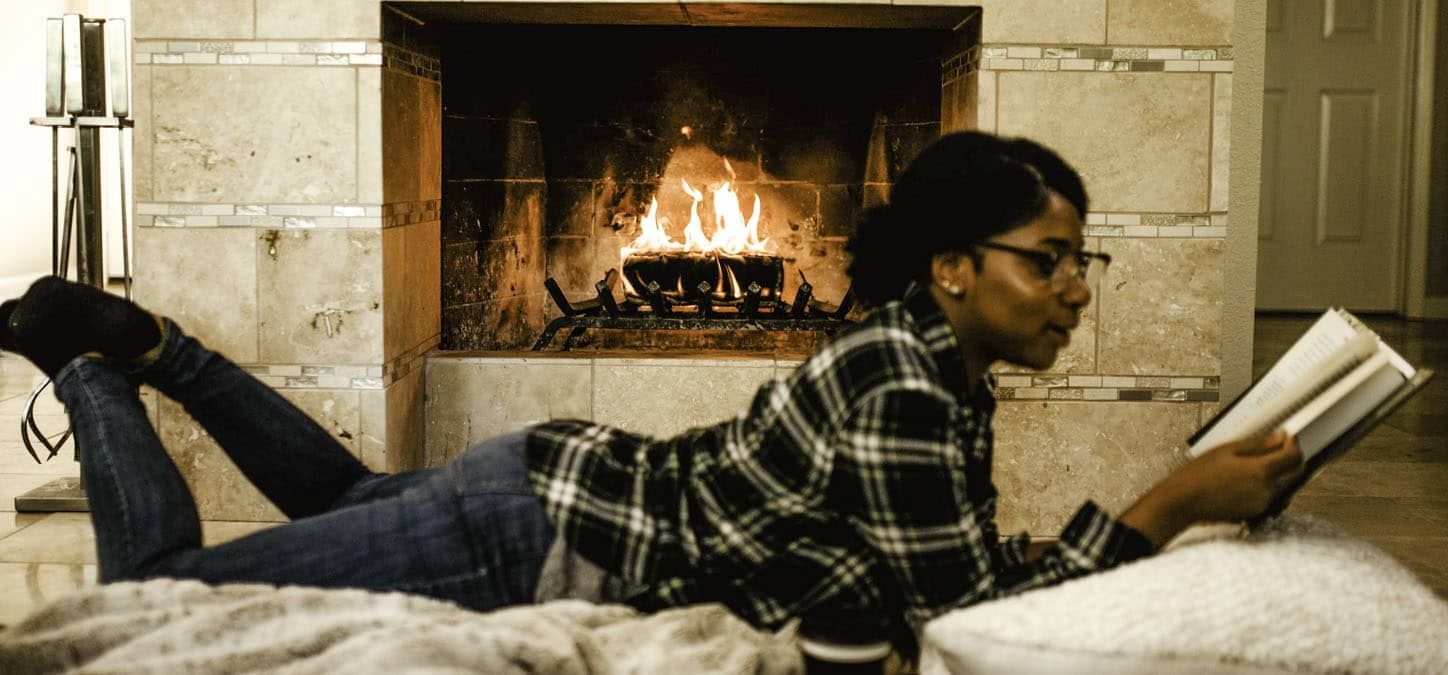A woman is laying on the floor reading a book in front of a fireplace.