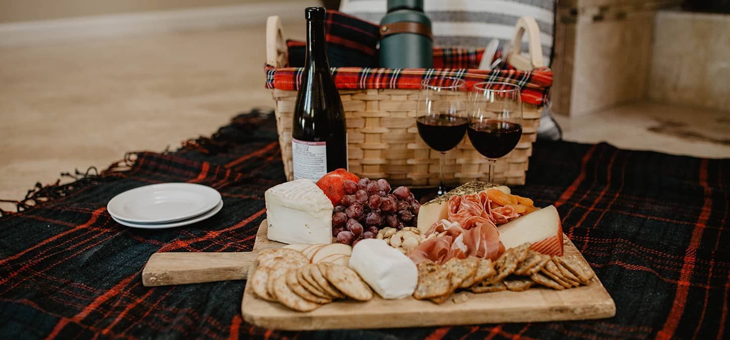 A picnic basket filled with wine , cheese , crackers and fruit is sitting on a blanket.