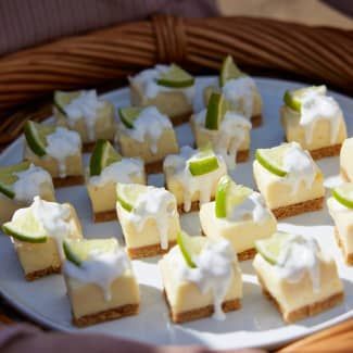 Mini key lime pie bites on a white plate, topped with cream and lime slices, in front of a woven basket.