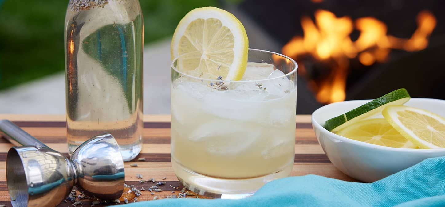 A glass of lavender gin sour with ice and a slice of lemon on a table with a fire pit in the background