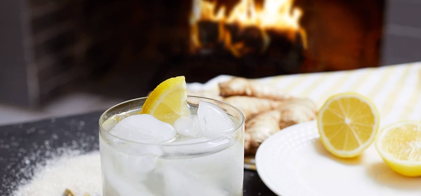 A glass of lemon ginger shrub mocktail with a lemon slice on a table in front of a fireplace.