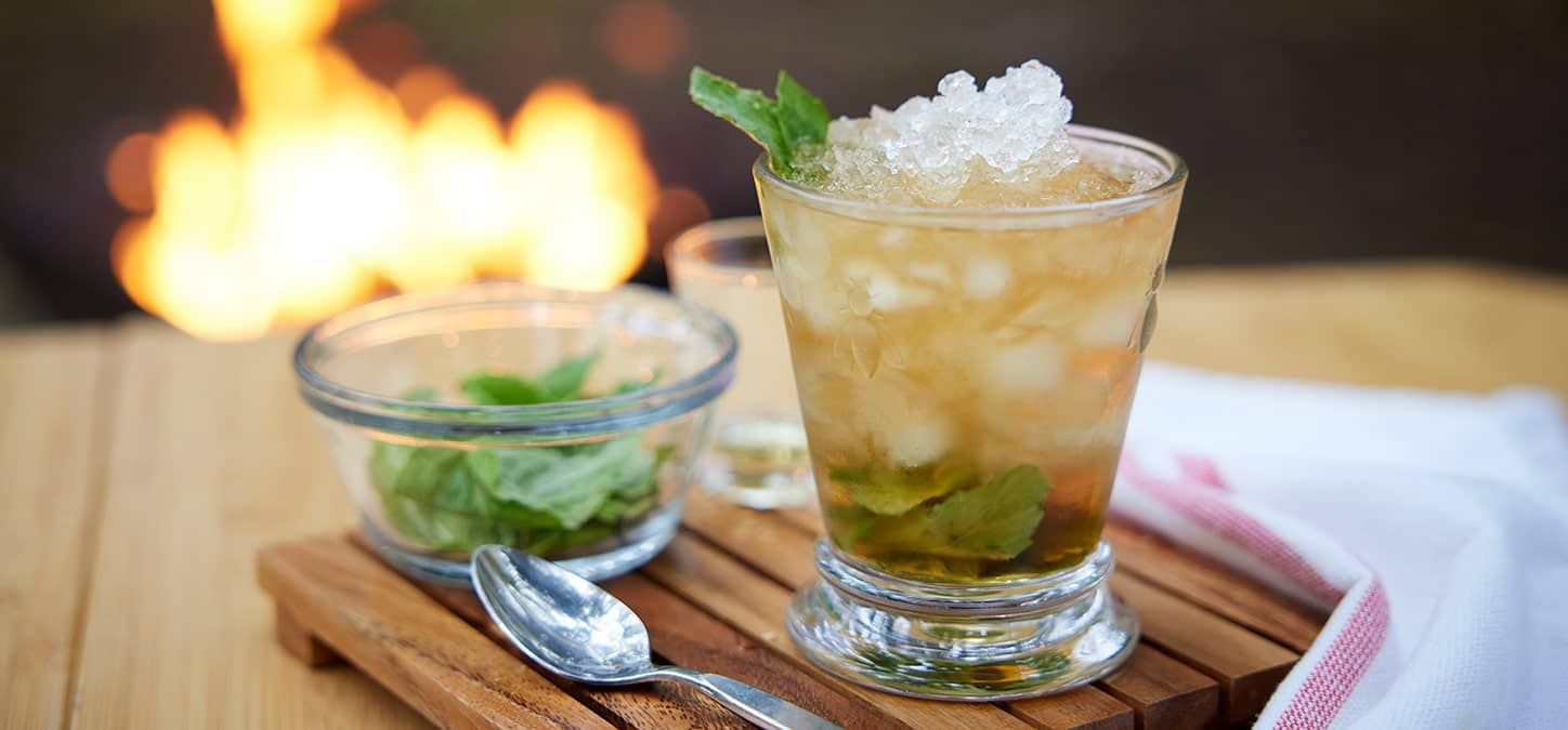 A glass of mint julep with mint leaves and a spoon on a wooden tray with a fire burning in the background