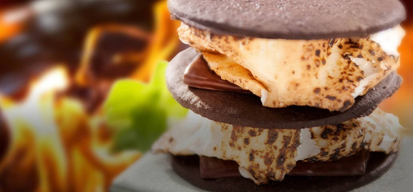 A s'mores sandwich stacked with marshmallows, chocolate mint and chocolate cookies with a fire in the background.