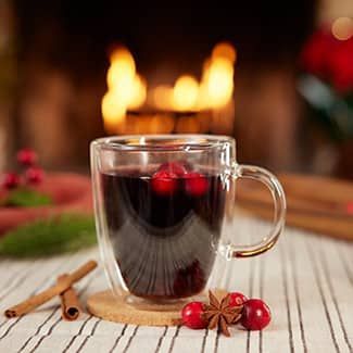 Mug of mulled wine with cranberries and cinnamon sticks in front of a fireplace.