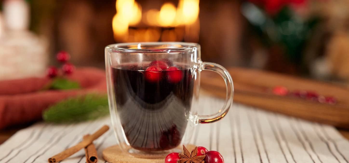 A cup of mulled wine with cranberries and cinnamon sticks on a table in front of a fireplace.