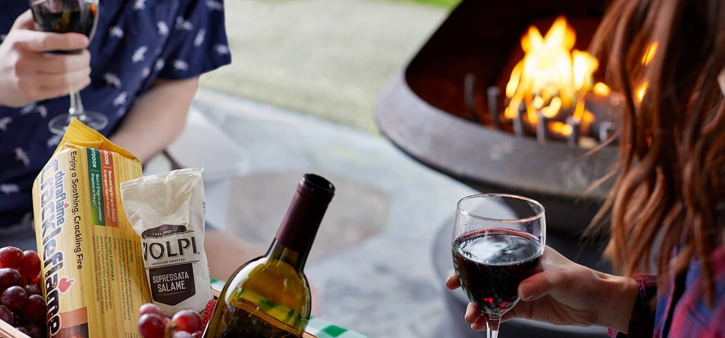 Two people are sitting at a table with a bottle of wine and a glass of wine outdoors with a fire pit